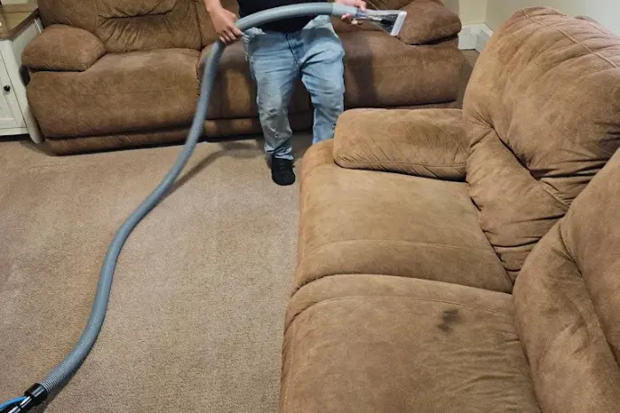 Technician cleaning beige carpet with a long hose between two brown fabric sofas in a family room